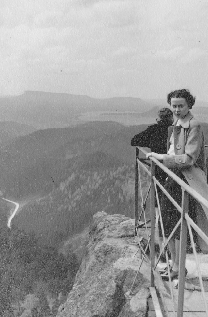 Hilde Nordhoffe steht auf einer Aussichtplattform auf einem Berg. Sie sieht in die Kamera, im Hintergrund ist eine weitere Person zu sehen. 