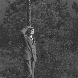 Roland Nordhoff lehnt an einen Baum, seine Hand zum Schutz vor das Gesicht haltend, schaut er in die Sonne. Im Hintergrund Nadelbäume..  