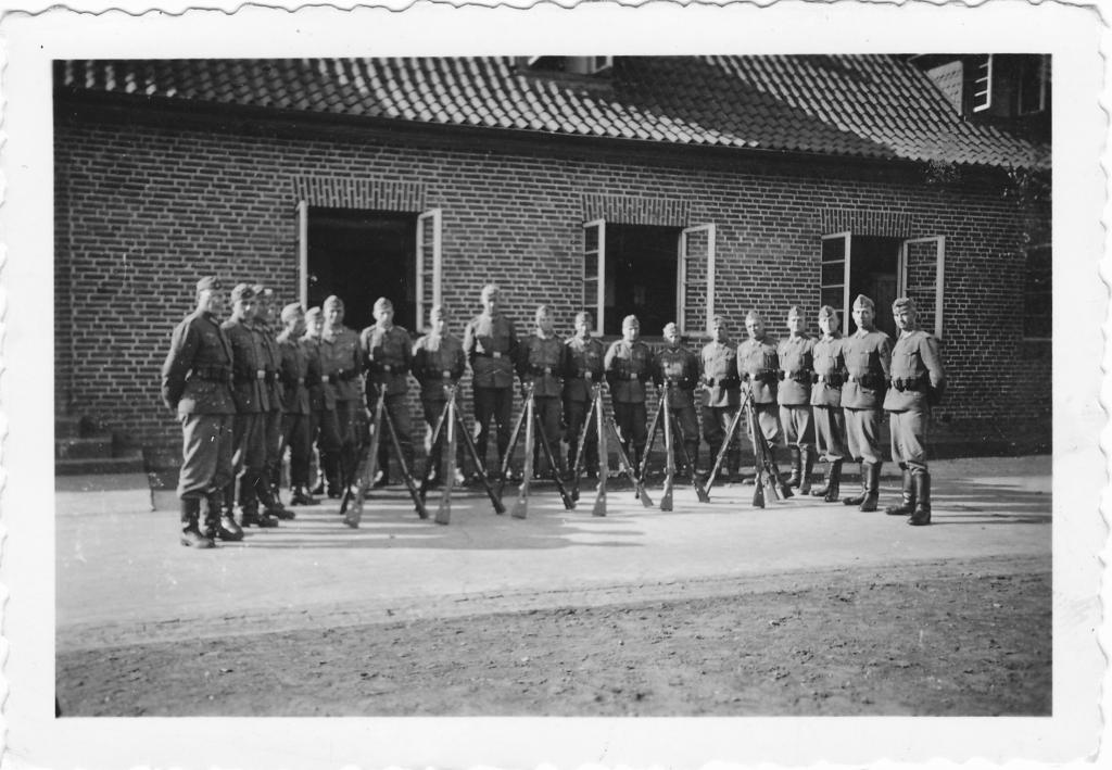 Eine Gruppe von 19 Wehrmachtsoldaten steht vor einem Gebäude, vor ihnen einige Gewehre in Ruheposition. 