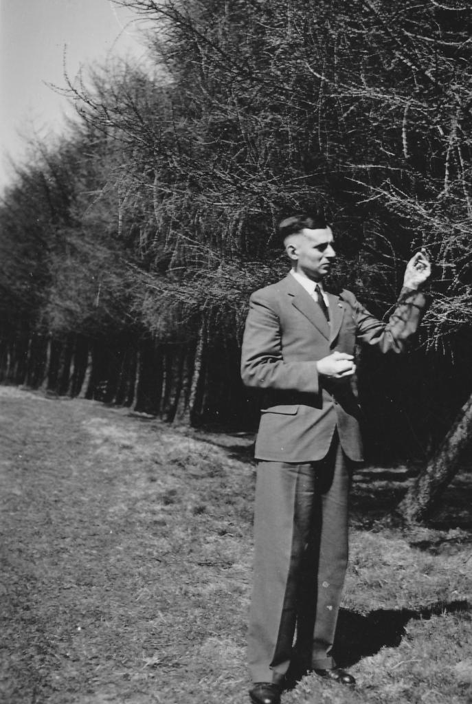 Roland Nordhoff steht vor einem Baum und fasst an einen Zweig, zu sehen von vorne, vom Knie bis zum Scheitel, im Anzug. Im Hintergrund dichte Bäume. 