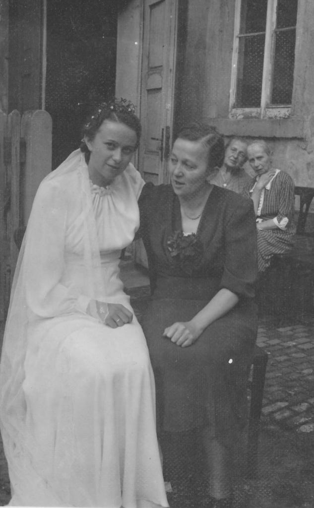 Hilde Nordhoff in einem weißen Hochzeitskleid und eine weitere Frau sitzen vor einem Gebäude. 