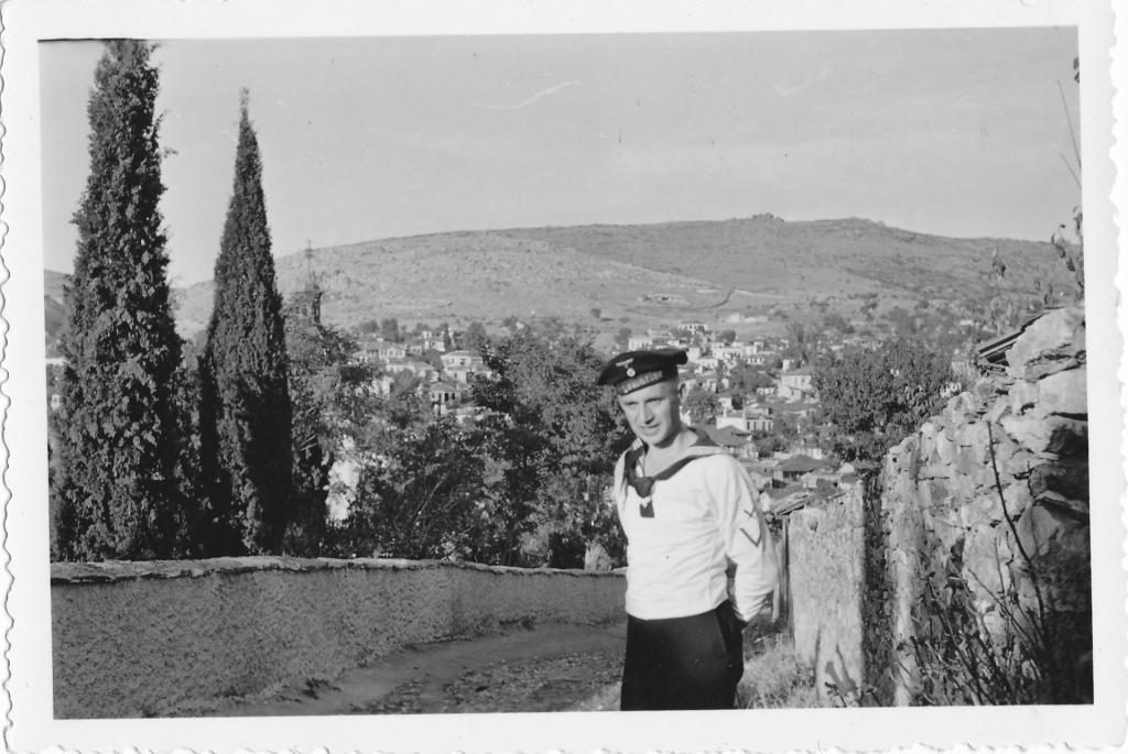 Roland Nordhoff in Marineuniform mit dunkelblauer Schirmmütze, der für die Kamera posiert. Im Hintergrund ein Ort am Hang eines Berges, am linken Bildrand befinden sich zwei Zypressen.