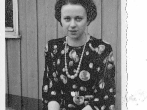 Halbnahe Aufnahme von Hilde Nordhoff, die vor einem Holzhaus sitzt. Sie trägt ein Kleid mit Punkten und eine große Perlenkette. 