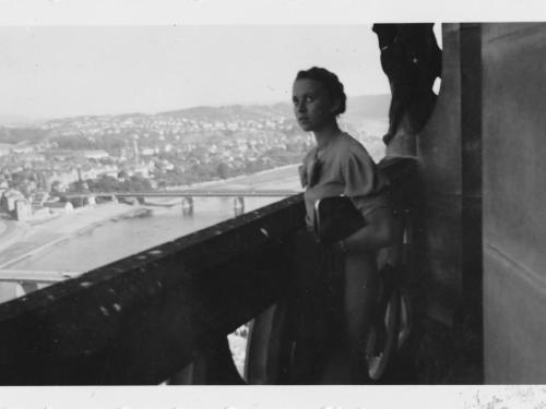 Halbnahe Aufnahme von Hilde Nordhoff, die an einer Ballustrade auf einem Turm lehnt. Im Hintergrund ist ein Fluss zu sehen, über den eine Brücke führt, und an den sich die Stadt Meißen schmiegt. 