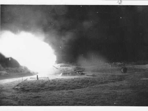 Umrisse eines militärischen Objektes, welches lichterloh in brand steht. Im Hintergrund sitzt eine Gruppe Soldaten. Nachtaufnahme.