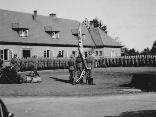 Rückenansicht dreier Männer in Wehrmachtuniform, in ihrer Mitte eine nationalsozialistische Flagge. Im Hintergrund stehen Wehrmachtsoldaten, wohl ein Zug, zum Appell vor einem Gebäude.
