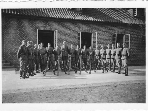 Eine Gruppe von 19 Wehrmachtsoldaten steht vor einem Gebäude, vor ihnen einige Gewehre in Ruheposition. 