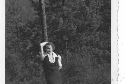 Hilde Nordhoff lehnt an einen Baum, mit dem rechten Arm hät sie sich über dem Kopf am Baumstamm fest. Im Hintergrund Nadelbäume.  