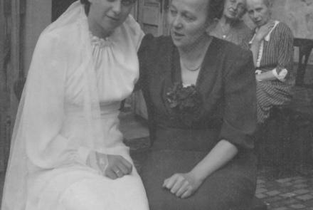 Hilde Nordhoff in einem weißen Hochzeitskleid und eine weitere Frau sitzen vor einem Gebäude. 